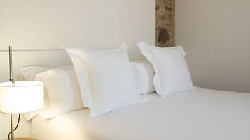 Una cama bien hecha con sábanas y almohadas blancas, una lámpara de noche sobre una mesita de madera, y una pared lisa con textura decorativa.