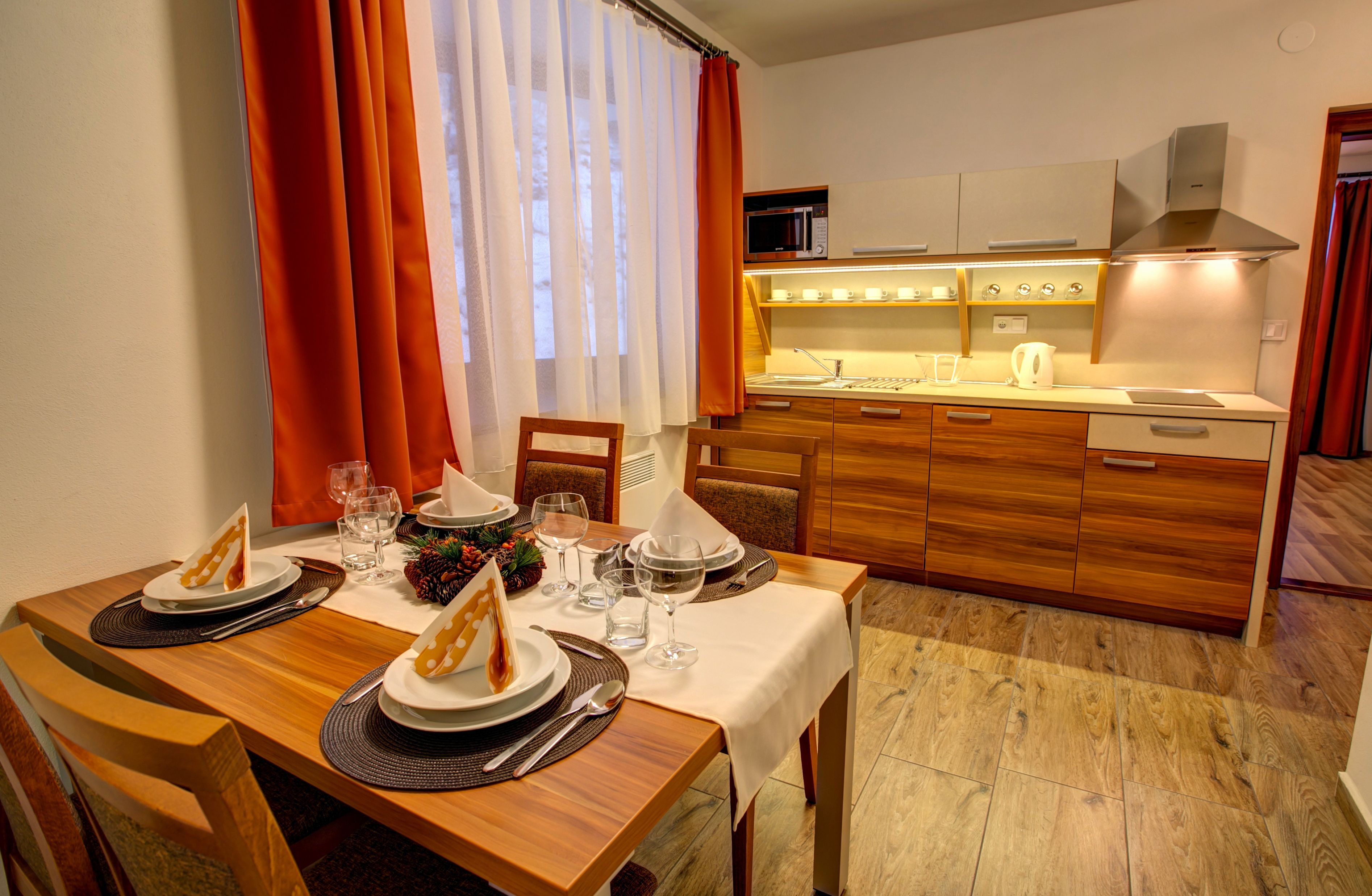 A dining area with a wooden table set for two, a kitchen with modern appliances, and a view of the dining area through a window with curtains.