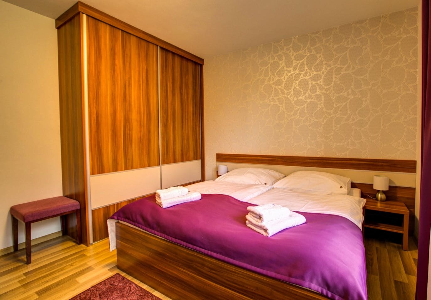 Un dormitorio con estructura de cama de madera, ropa de cama púrpura, una mesita de noche de madera, un banco y un papel tapiz estampado.