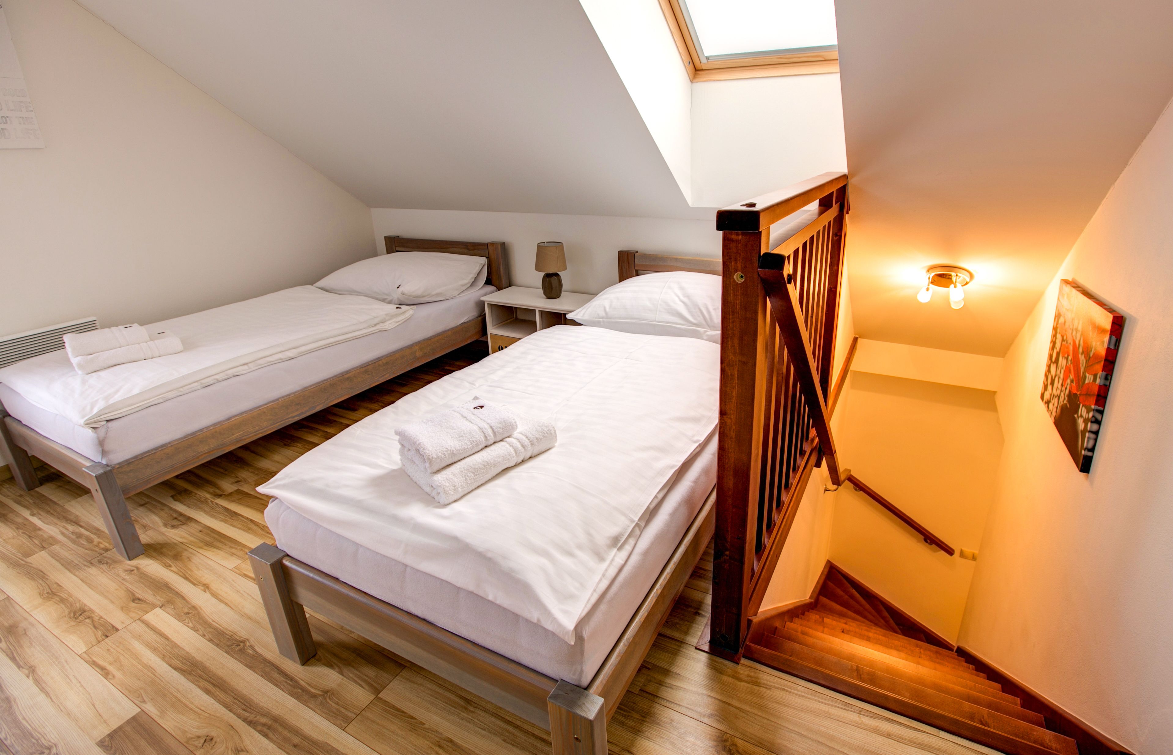 A bedroom with two single beds, wooden flooring, a skylight, a staircase, and a wall-mounted light fixture.