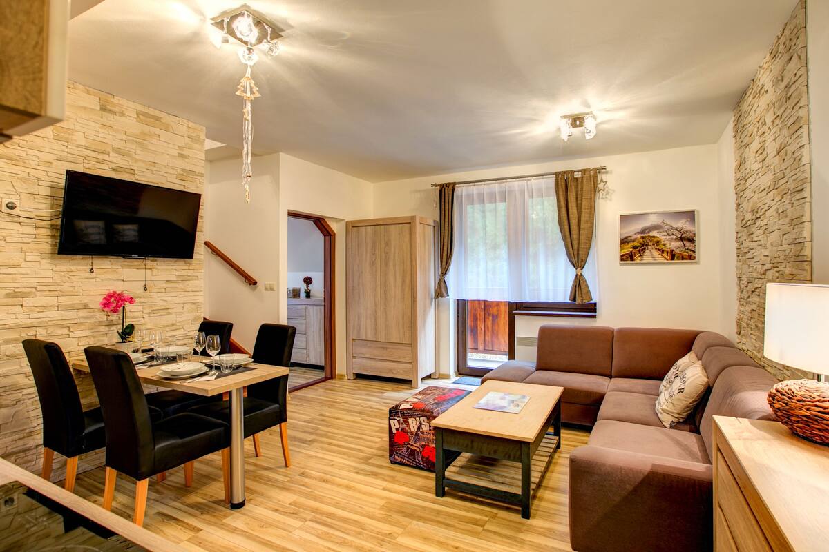 Ein modernes Wohnzimmer mit Essbereich, einer Couch, einem an einer Steinwand befestigten Fernseher und einer Treppe, die in einen anderen Raum führt.