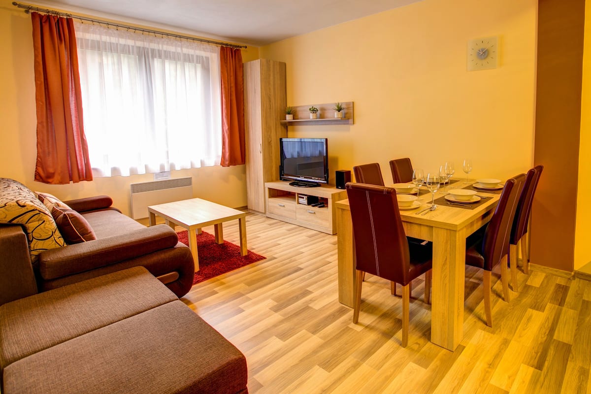 Ein modernes Wohnzimmer mit einer Couch, einem Esstisch mit Stühlen, einem Fernseher und einem kleinen Regal mit Pflanzen.