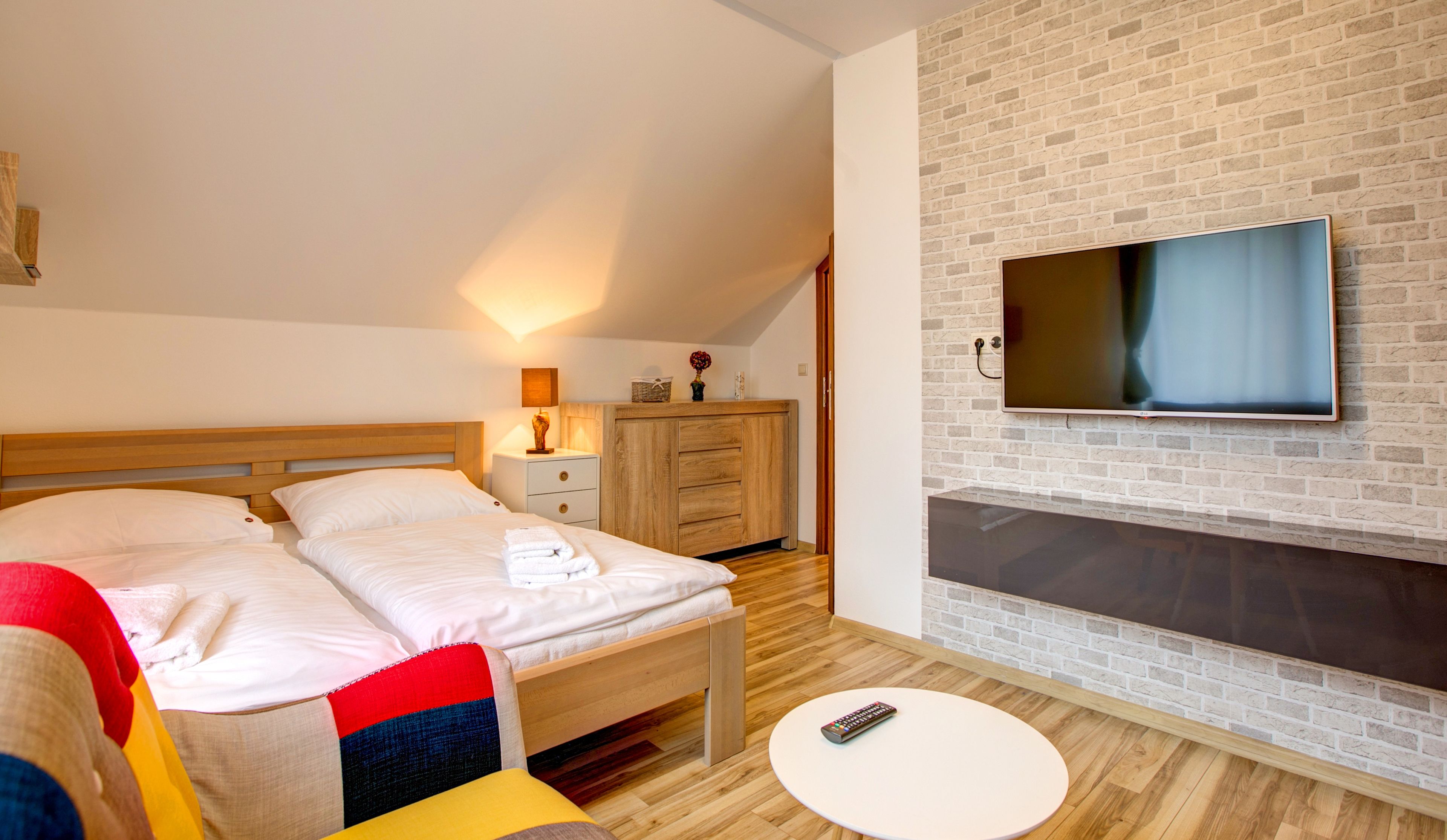 A hotel room with a bed, a TV mounted on a brick wall, a bedside table with a lamp, and a small round table with a remote control.
