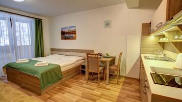 Quarto de hotel com cama, escrivaninha, cadeira, pequeno espaço de cozinha e janela com cortinas.