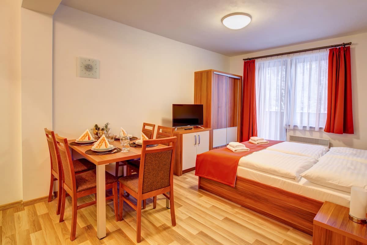 Ein Hotelzimmer mit einem Holzespeisetisch, gedeckt für zwei Personen, einem Fernseher auf einer Holzkommode, einem Bett mit roter Bettwäsche und einem Fenster mit roten Vorhängen.