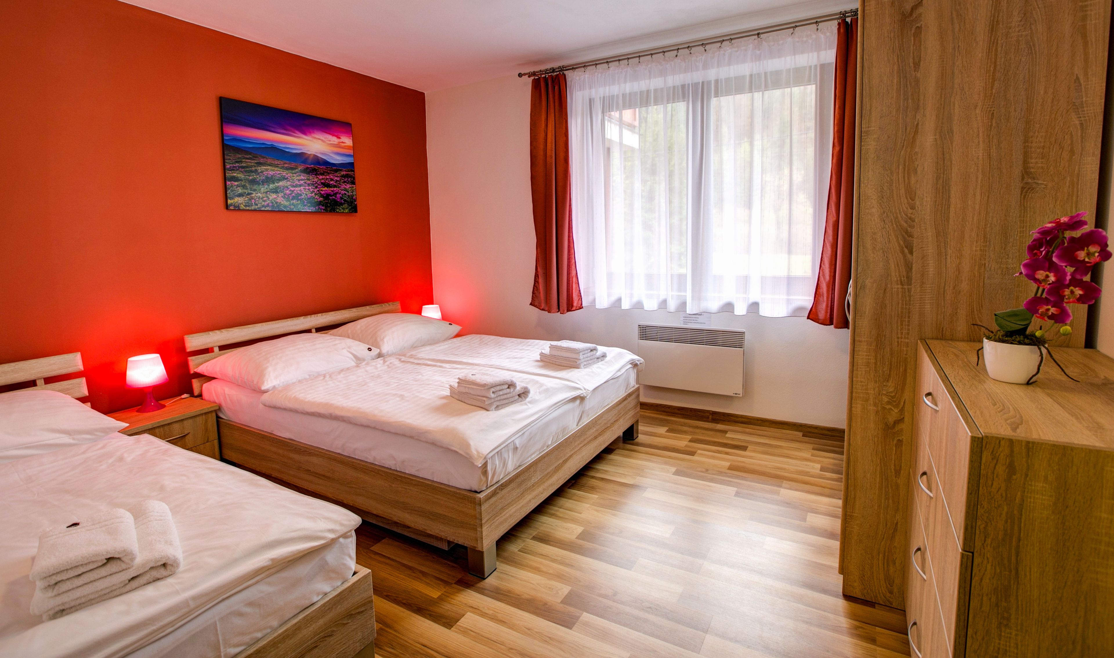 A hotel room with two beds, a painting on the wall, a window with curtains, and a potted orchid on a wooden dresser.
