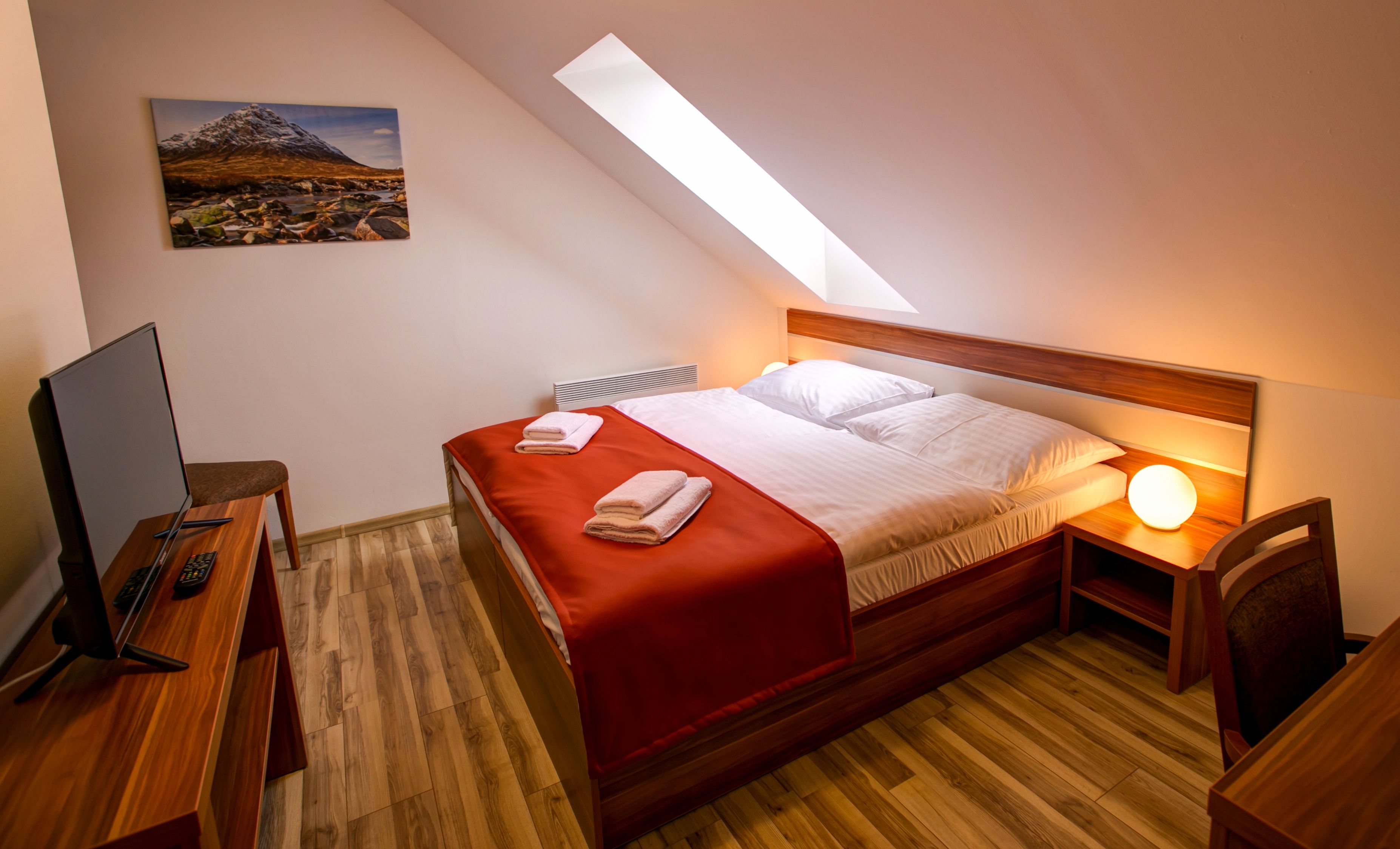 Um quarto com teto inclinado, uma cama com roupa de cama branca, um cobertor vermelho, uma escrivaninha com um computador e um quadro na parede.