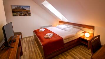 Um quarto com teto inclinado, uma cama com roupa de cama branca, um cobertor vermelho, uma escrivaninha com um computador e um quadro na parede.