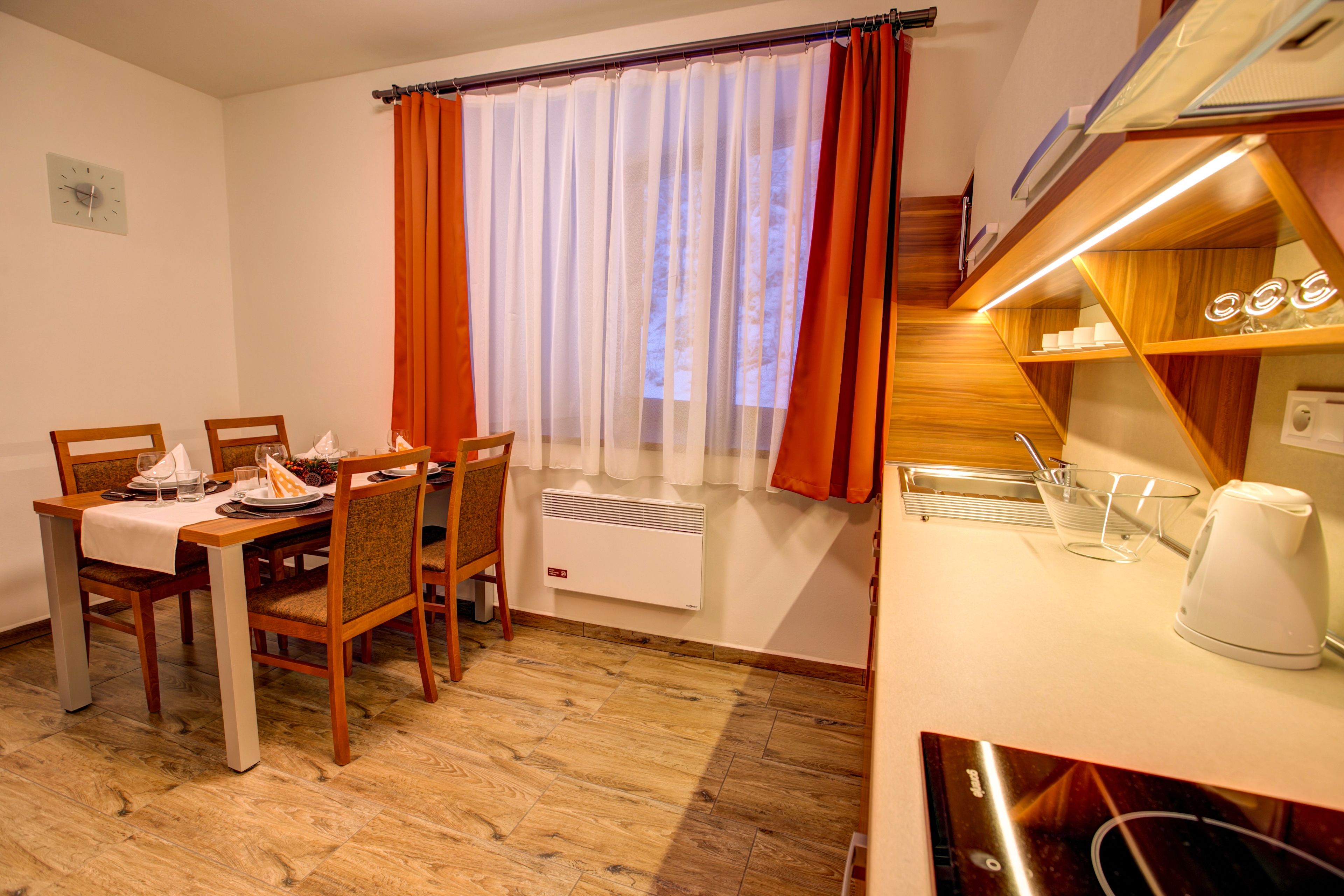Een eetgedeelte met een tafel voor vier personen, een raam met oranje gordijnen, een witte radiator, en een keuken met een waterkoker en planken.