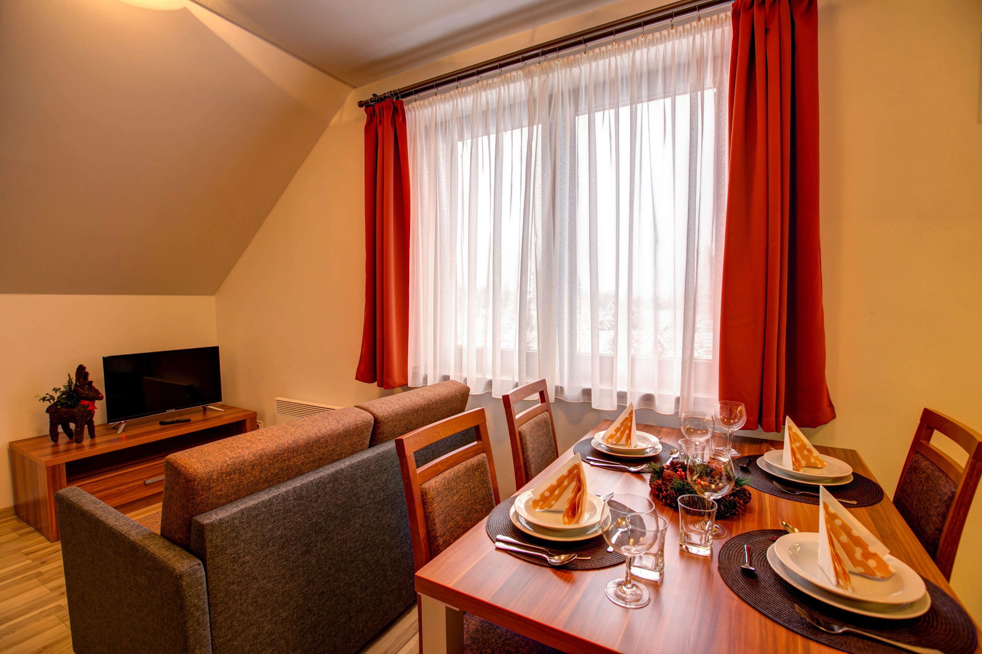 A dining area with a wooden table set for two, a television on a wooden stand, and a sofa in the background.