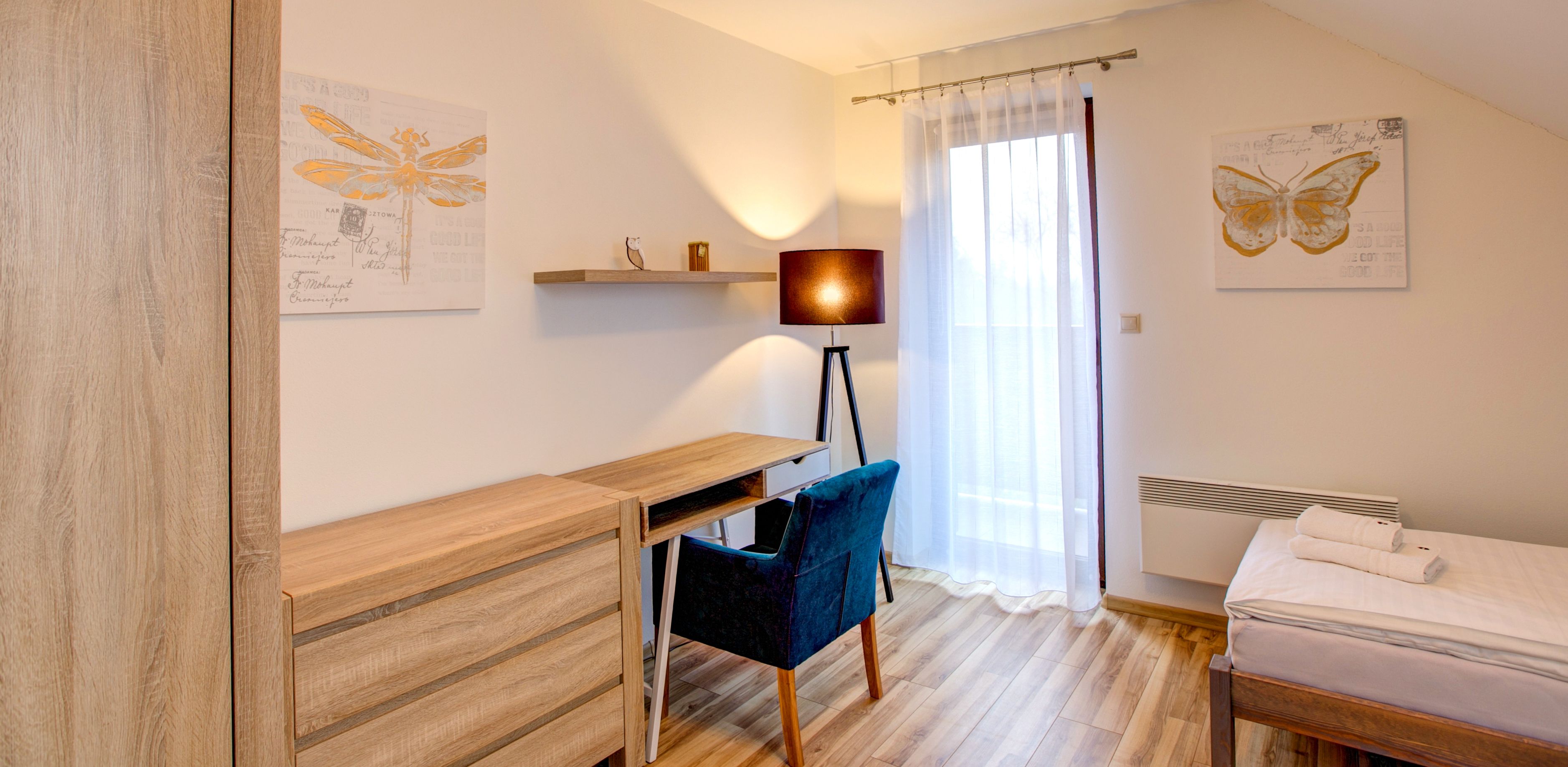 A bedroom with a wooden desk, a chair, a bed with a white duvet, a standing lamp, and two butterfly artwork on the walls.