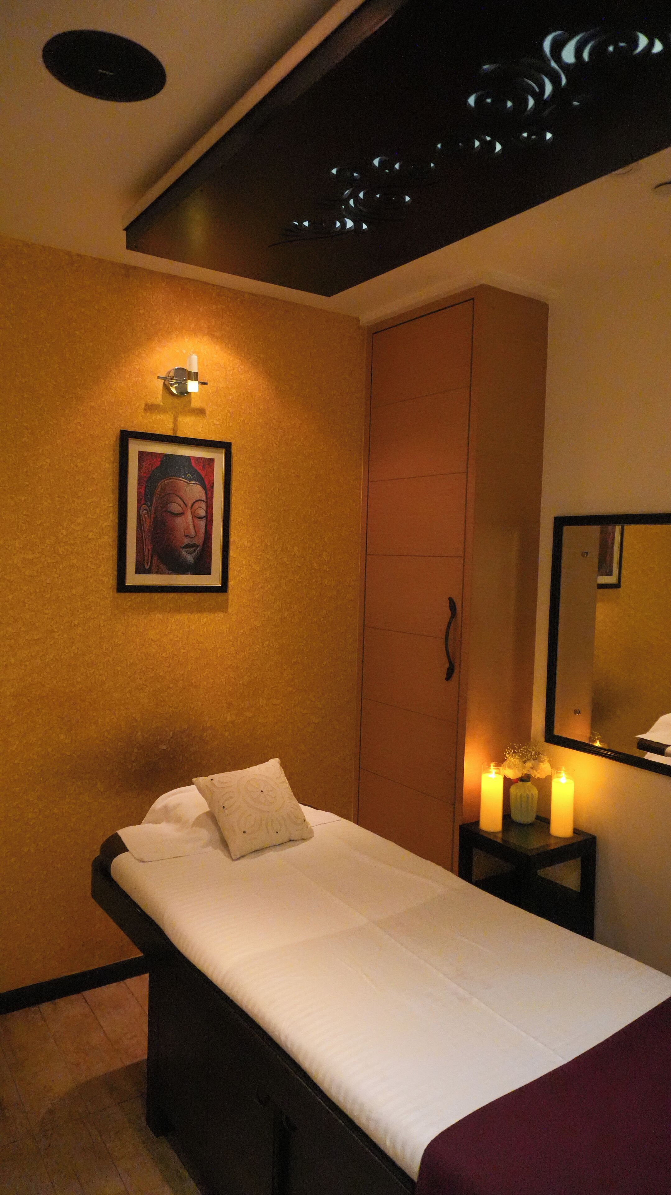 A single bed with a white mattress and a decorative pillow, a wall-mounted light fixture, a framed artwork, a mirror, and a small table with candles.