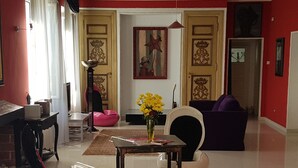 Ein Wohnzimmer mit einem Klavier, einem pinken Sitzsack, einem violetten Sofa und einer Vase mit gelben Blumen.