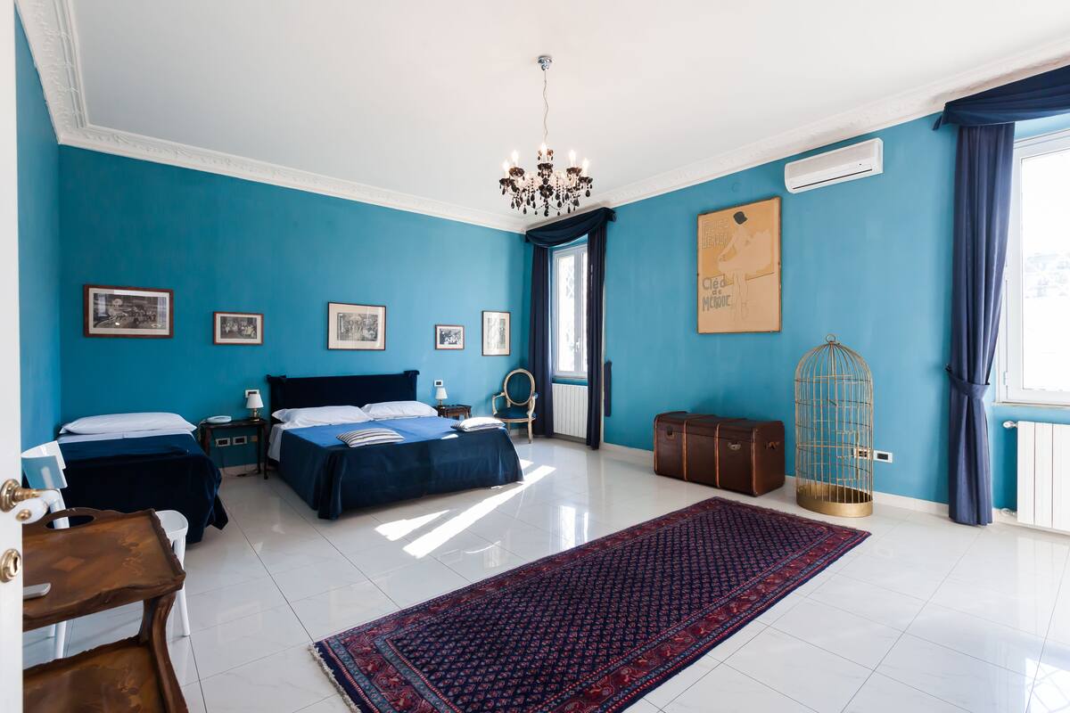A bedroom with two beds, a chandelier, a large window with curtains, a radiator, and a birdcage.