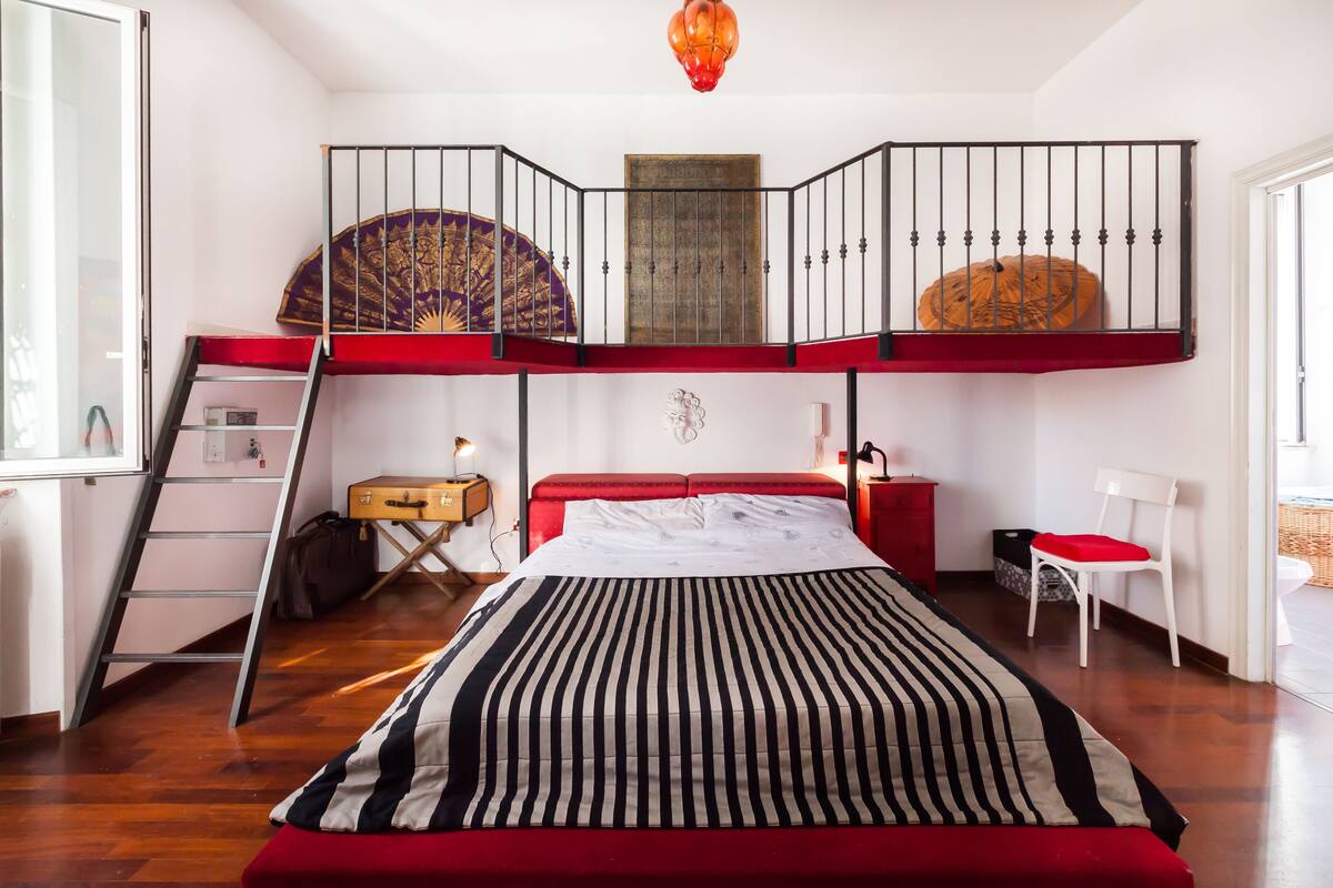 A bedroom with a bunk bed, a ladder, a red headboard, a bedside table, a chair, and a window.