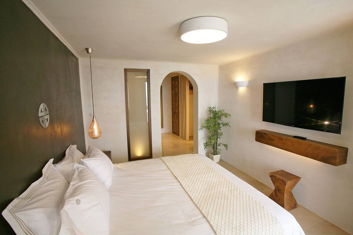 A modern hotel room with a large bed, a flat-screen TV mounted on the wall, a bedside table, and a mirror.