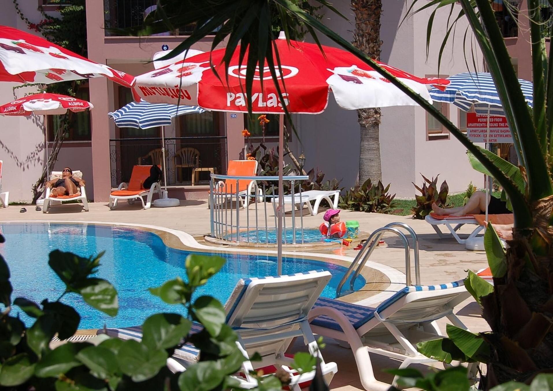 A pool area with lounge chairs and umbrellas, a lifeguard stand, and a building in the background.
