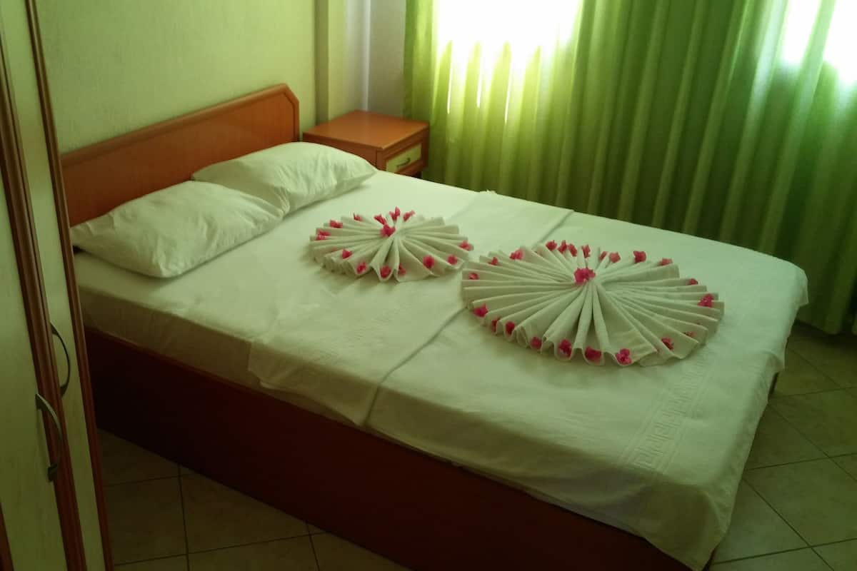 A neatly made bed with white linens and decorative pillows, a wooden headboard, a small wooden nightstand with a drawer, and a green curtain in the background.