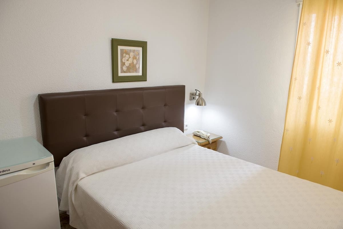 Une chambre d’hôtel comprenant un lit, une table de chevet avec une lampe, un téléphone et un petit bureau avec une imprimante.
