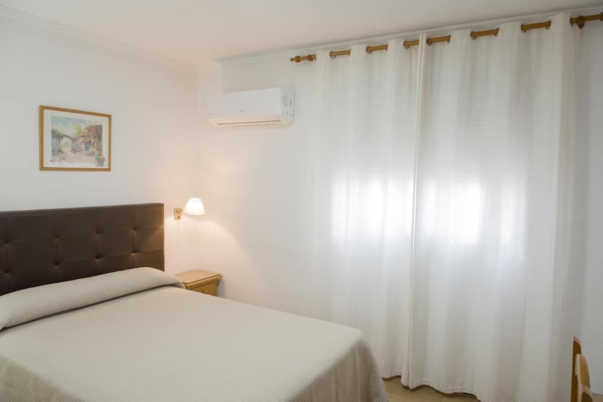 Une chambre d’hôtel comprenant un lit, une table de chevet, une lampe, un appareil de climatisation et une fenêtre avec des rideaux.