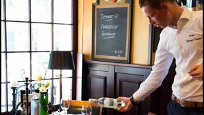 A man pouring a drink at a restaurant table with a chalkboard menu, salt and pepper shakers, and a vase with flowers. - Hotel Duhoux Leeuwarden (Wirdum)