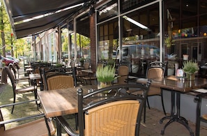 An outdoor cafe with wicker chairs and tables, large windows, and a covered seating area. - Hotel Duhoux Leeuwarden (Wirdum)