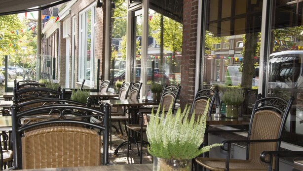 An outdoor cafe with wicker chairs and tables, large windows, and potted plants. - Hotel Duhoux Leeuwarden (Wirdum)