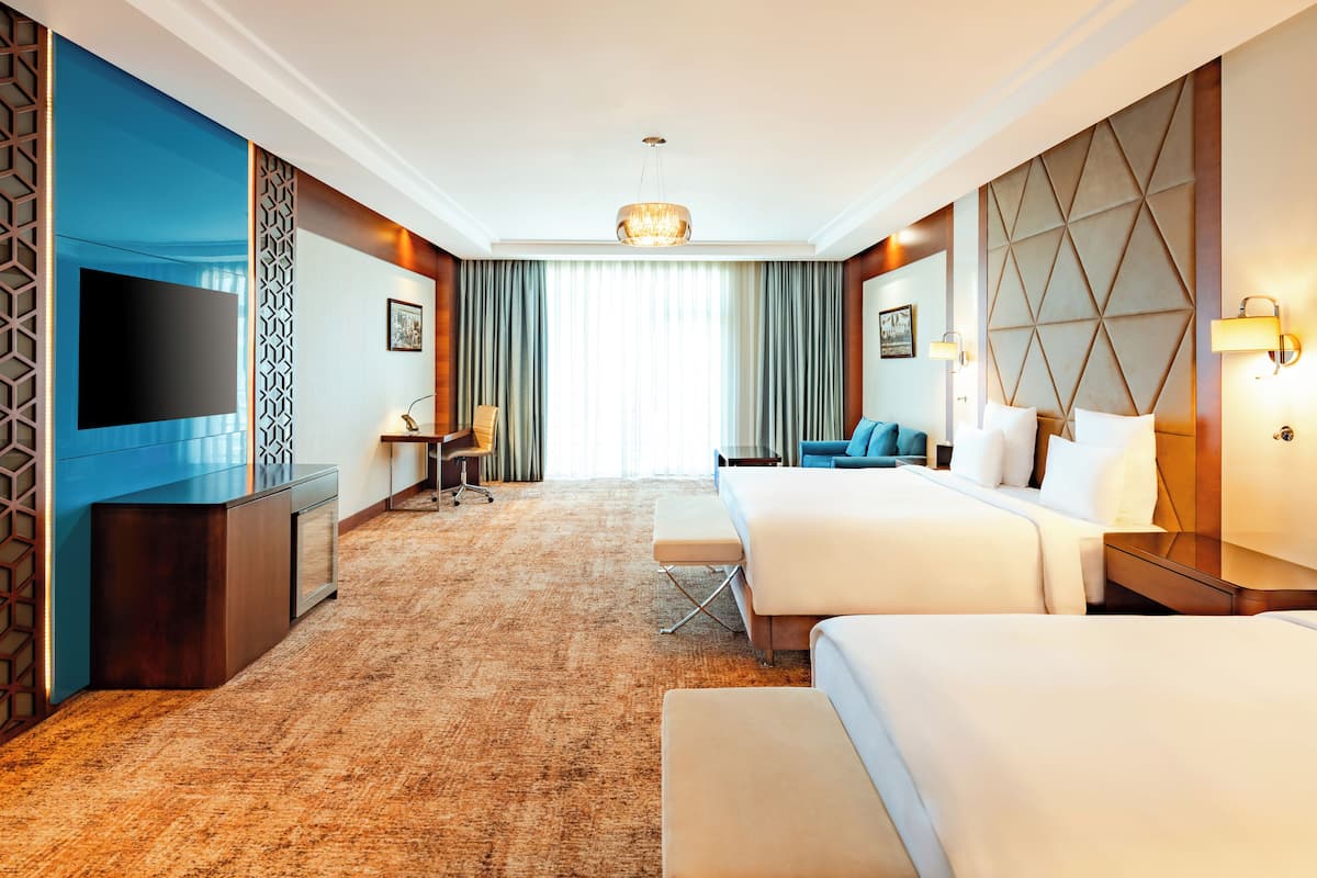 Ein Hotelzimmer mit großem Flachbildfernseher, Schreibtisch mit Stuhl, zwei Betten, einer Couch und einem großen Fenster mit Vorhängen.