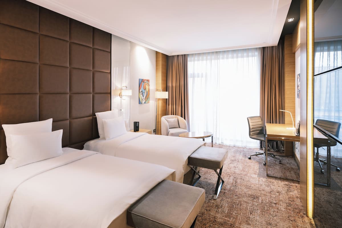 Ein Hotelzimmer mit zwei Betten, einem Schreibtisch, einem Stuhl und einem großen Fenster mit Vorhängen.