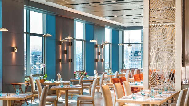 Un restaurant moderne avec de hauts plafonds, de grandes fenêtres et une vue sur la ville. Les tables sont disposées avec soin et des chaises sont placées autour, tandis que des luminaires suspendus illuminent l’espace.