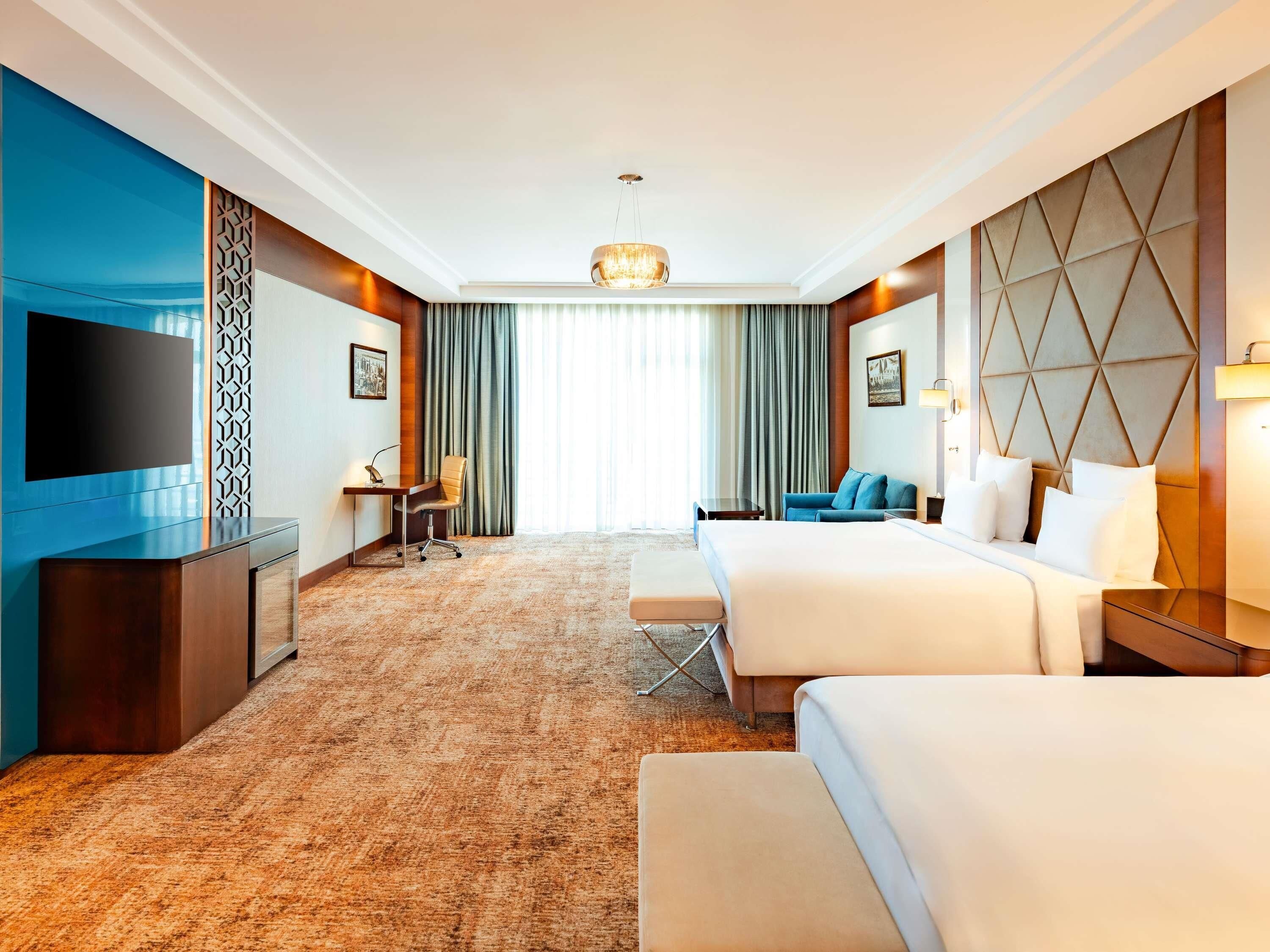 Une chambre d’hôtel avec un grand lit, un lit plus petit, un bureau avec une chaise, une télévision et une grande fenêtre avec des rideaux.