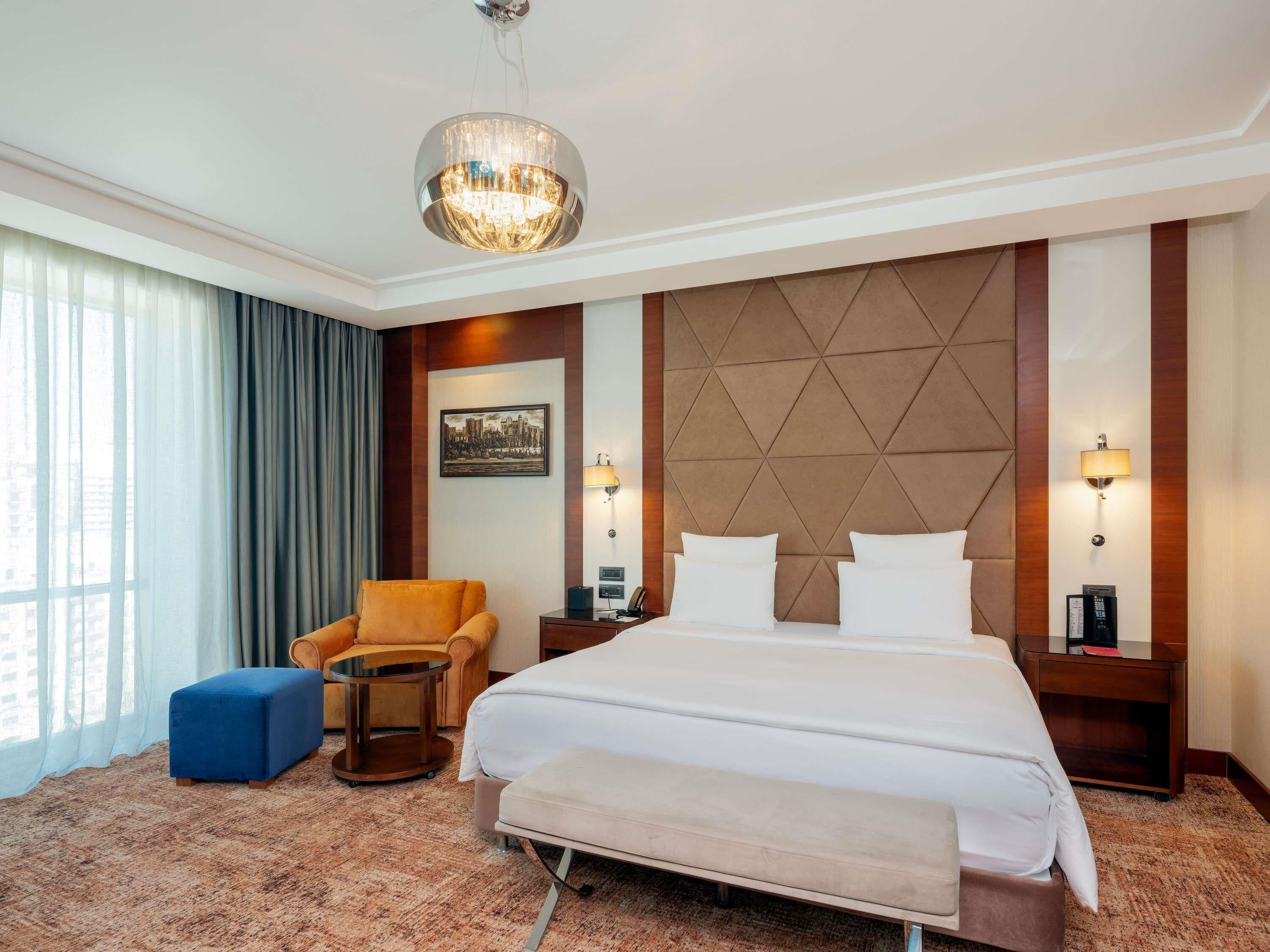 A hotel room with a large bed, two bedside tables, a chair, a small table, and a large window with curtains.