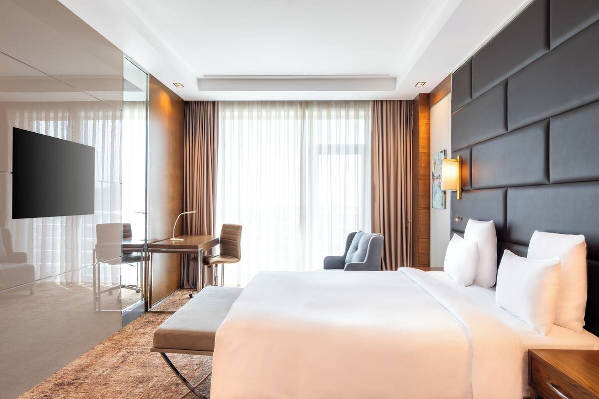 Ein Hotelzimmer mit einem großen Bett, einem Schreibtisch mit Stuhl, einem Fernseher und einem großen Fenster mit Vorhängen.