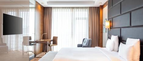 A hotel room with a large bed, a desk with a chair, a TV, and a large window with curtains.
