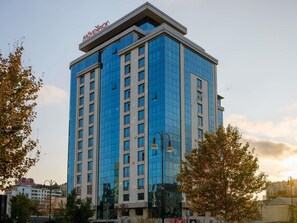 Ein modernes Hochhaus mit einer reflektierenden Glasfassade, einer gut sichtbaren Beschilderung obenauf und einer umliegenden Stadtlandschaft mit Bäumen und weiteren Gebäuden.