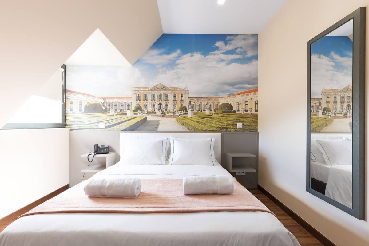 Une chambre d’hôtel avec un grand lit, un bureau et une vue sur un bâtiment historique, visible à travers une lucarne.