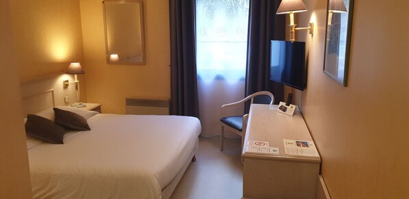 Habitación de hotel con cama, escritorio, silla, televisor y ventana con cortinas.