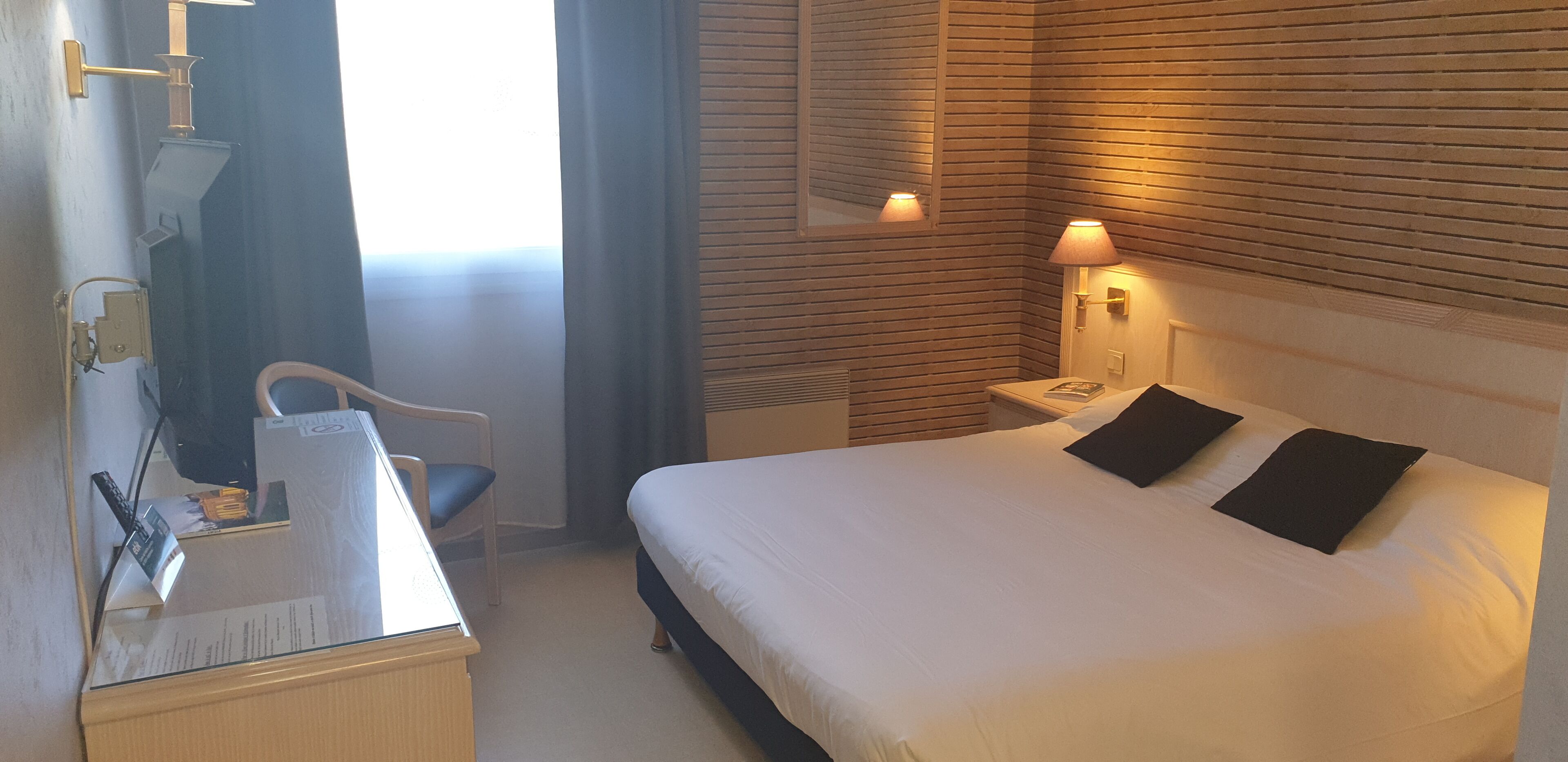 Una habitación de hotel con cama, lámpara de noche, escritorio, silla y ventana con cortinas.
