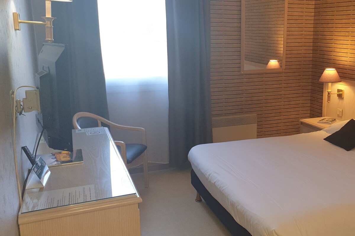 Une chambre d’hôtel avec un lit, une lampe de chevet, un bureau, une chaise et une fenêtre avec des rideaux.