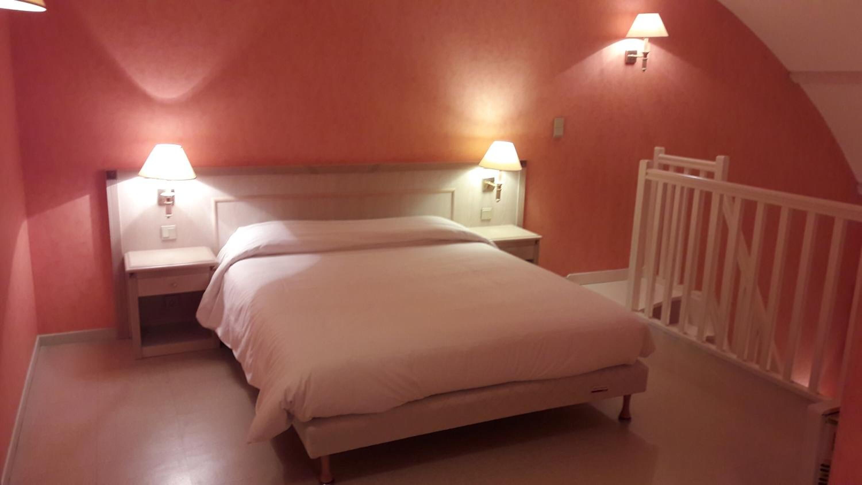 Un lit simple dans une chambre avec deux tables de chevet, chacune équipée d’une lampe. Les murs sont peints dans une couleur chaude, et une rambarde blanche se trouve sur le côté droit.