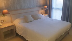 Un llit ben fet amb llençols blancs, dues làmpades de nit, una tauleta de nit amb un telèfon i una cadira.