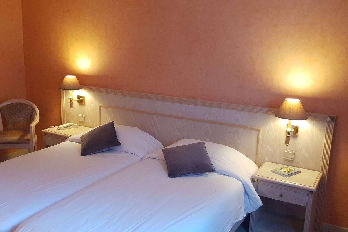Habitació d'hotel amb un llit gran, dues làmpades de nit, una cadira i una tocador amb un llibre.