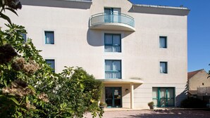 Un edifici de diversos pisos amb un balcó corbat al segon pis, una entrada central amb una porta i una petita zona de jardí al davant.
