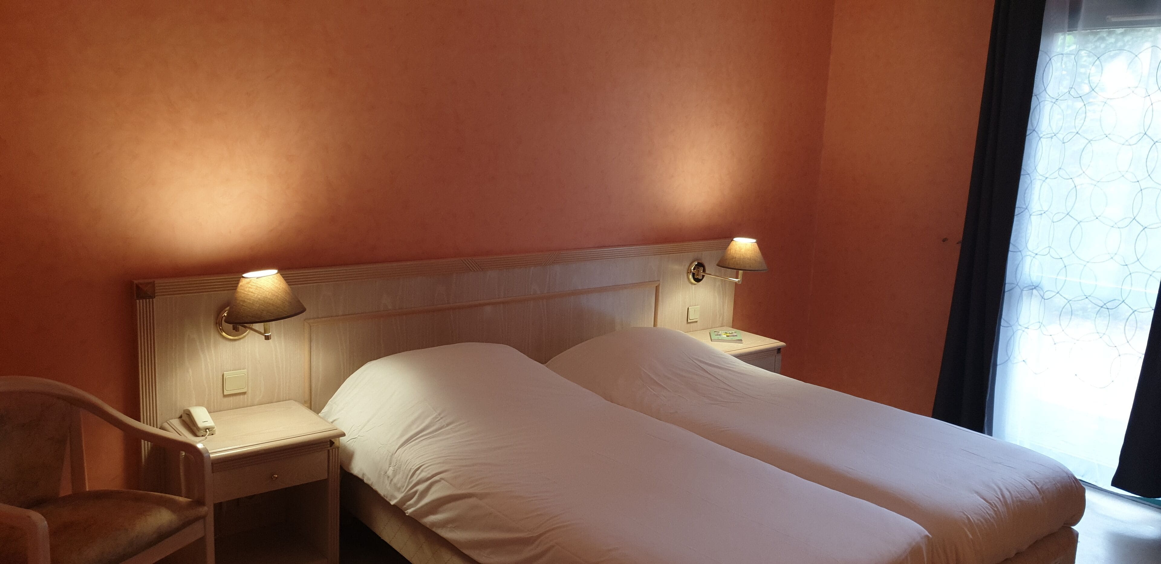 A hotel room with two beds, a nightstand, a lamp, and a chair.