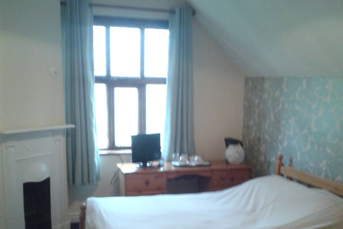 A bedroom with a bed, a desk, a chair, a window with curtains, and a radiator.