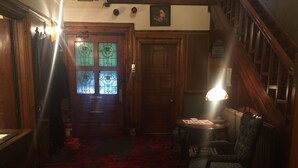 A dimly lit room with a wooden staircase, a door with stained glass windows, and a table with a lamp.