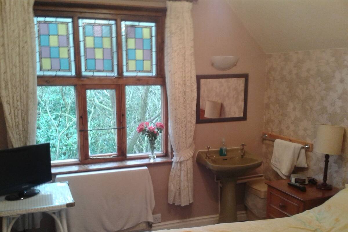 A bedroom with a bed, a bedside table with a lamp, a small desk with a TV, a window with curtains, and a bathroom with a sink and mirror.