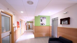 A hospital corridor with wooden doors, a reception desk, a waiting area with a couch, and informational posters on the walls.