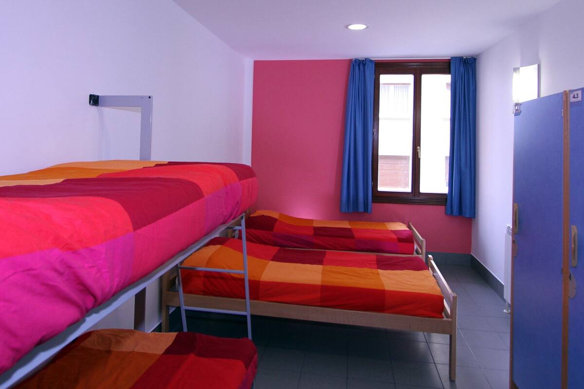 A dormitory room with bunk beds, a window with blue curtains, and a door with a number.