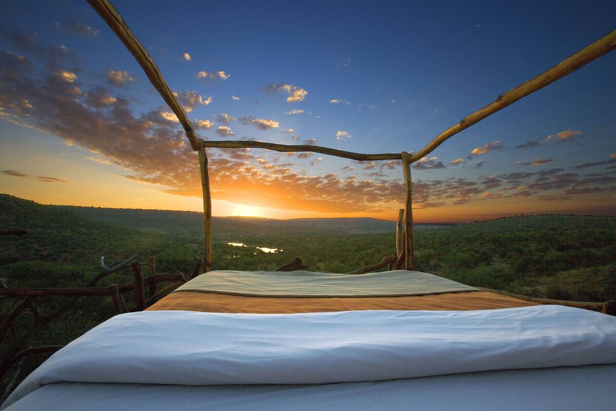 A bed with a view of a sunset over a hilly landscape.
